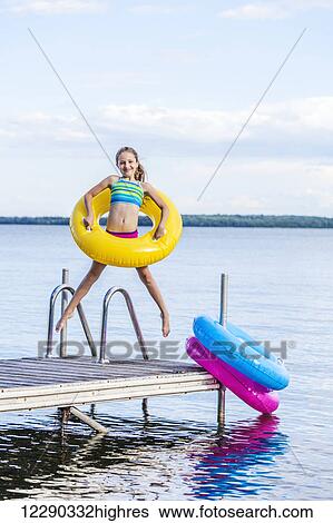 Stock Photograph - Girl jumping on dock by Balsam Lake holding a yellow inflatable ring; Ontario, Canada. Fotosearch