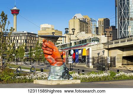 Stock Photograph - Large hand sculpture holding a metal (paper) plane, with commuter train, city buildings and the Calgary Tower in background with blue sky; Calgary, Alberta, Canada. Fotosearch