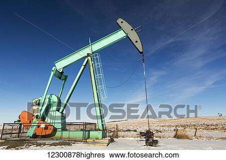 Stock Photograph - Large pump jack in a snow covered field with snow covered foothills, blue sky and clouds in the background; Longview, Alberta, Canada. Fotosearch