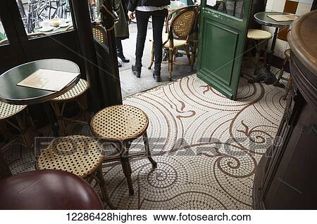 Stock Photograph - Looking down at the floor of a parisian cafe with small square tiles, traditional tables and stools, and the wooden bar; Paris, France. Fotosearch