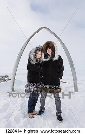 Stock Photograph - Male and female native youths wear traditional fur parkas while standing next to the Browerville Walebone arch, Barrow, North Slope, Arctic Alaska, USA, Winter. Fotosearch
