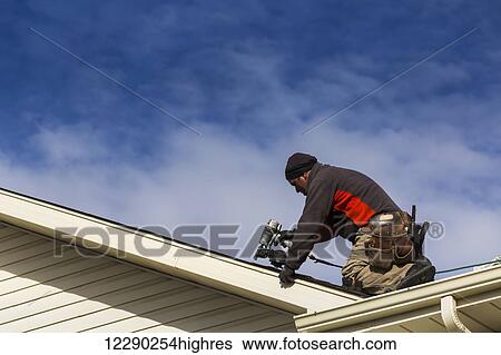 Stock Photograph - Male roofer with air nailer on top of roof nailing new shingles with clouds and blue sky; Calgary, Alberta, Canada. Fotosearch