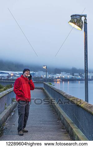 Stock Photograph - Man on a smart phone and standing on a boardwalk pier on a foggy evening, Ketchikan, Southeast Alaska, USA, Spring. Fotosearch