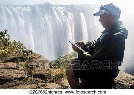 Stock Photograph - Man on smart phone in front of Victoria Falls waterfall; Victoria Falls, Zimbabwe. Fotosearch