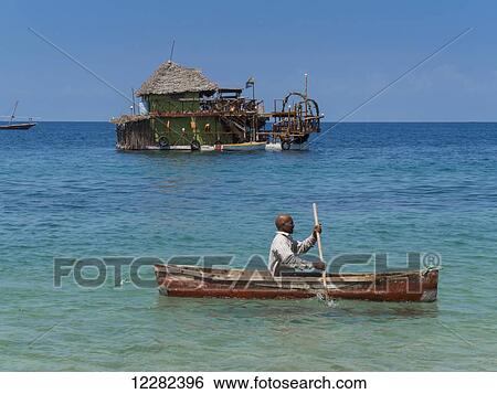 Stock Photograph - Man rowing in a boat past a floating building; Zanzibar, Tanzania. Fotosearch