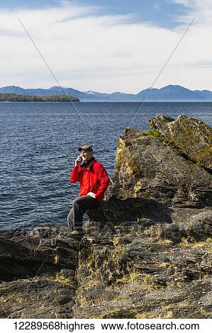 Stock Photograph - Man talks on his smart phone along the rocky shores of Tongass Narrows, Ketchikan, Southeast Alaska, Spring. Fotosearch