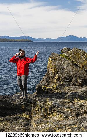 Stock Photograph - Man talks on his smart phone along the rocky shores of Tongass Narrows, Ketchikan, Southeast Alaska, Spring. Fotosearch