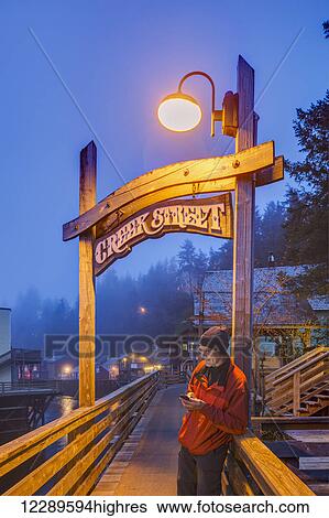 Stock Photograph - Man texting on a cell phone near the Creek Street sign and boardwalk on a foggy evening, downtown Ketchikan, Southeast Alaska, USA, Spring, HDR. Fotosearch
