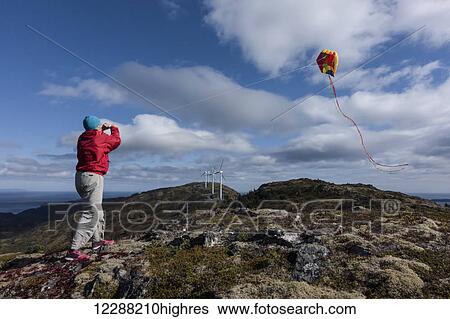 Stock Photograph - Mature woman in red jacket flying a kite on Pillar Mountain, Kodiak, Alaska with wind turbines in background, fall. Fotosearch