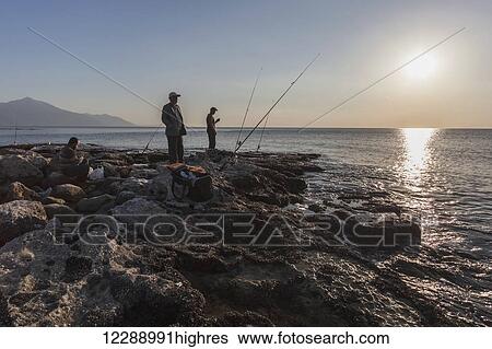 Stock Photograph - Men fishing at dusk at the water's edge of the Mediterranean, Mount Casius in the distance; Cevlik, Turkey. Fotosearch