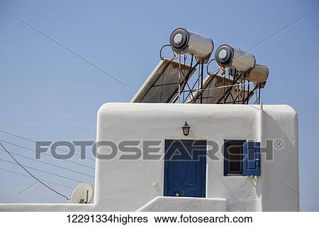 Stock Photograph - Modern solar panel powered hot water heaters for energy efficiency are mounted on the roof of a traditional greek home; Mykonos, Greece. Fotosearch