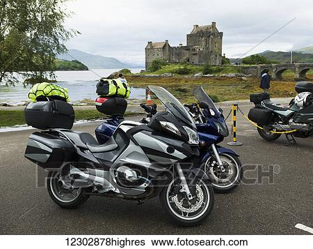 Stock Photograph - Motorcycles packed for a road trip and parked by the coast with a view of Eilean Donan Castle in the background; Scotland. Fotosearch