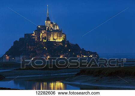 Stock Photograph - Night image of large stone Abbey perched on rocky island at low tide lit with flood lights with reflection in water; Mont St. Michel, Brittany, France. Fotosearch