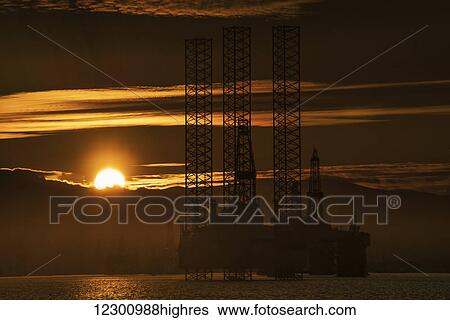Stock Photograph - Oil drilling rigs out in the ocean with a view of the coastline and golden sunset; Cromarty, Invergordon, Scotland. Fotosearch