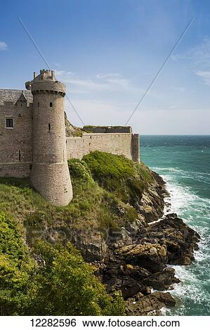 Stock Photograph - Old stone walled fort with turret on rocky shoreline along the sea with blue sky and clouds; Plevenon, Brittany, France. Fotosearch