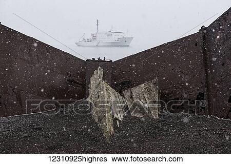 Stock Photograph - Old wooden boat abandoned on the shore at a whaling station with a cruise ship out in the water during a snow storm; Deception Island, South Shetlands, Antarctica. Fotosearch