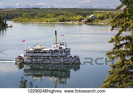 Stock Photograph - Paddlewheel boat on lake with tree lined shoreline, hills and mountains in the background and blue sky and clouds; Calgary, Alberta, Canada. Fotosearch