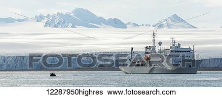 Stock Photograph - Panorama of a rigid inflatable and white ship in front of an ice cliff at the base of an Arctic glacier stretching back to mountains on the horizon; Spitsbergen, Svalbard, Norway. Fotosearch