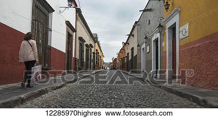 Stock Photograph - Pedestrian walking beside a cobblestone street and colourful buildings; San Miguel de Allende, Guanajuato, Mexico. Fotosearch