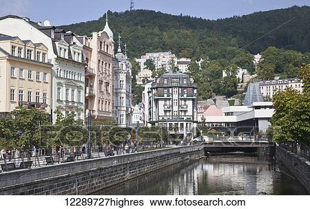 Stock Photograph - Pedestrians on a promenade along a river and residential buildings with a forested hillside; Karlova Vary, Czech Republic. Fotosearch