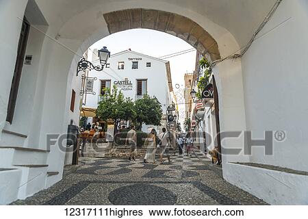 Stock Photograph - Pedestrians walking among buildings in Altea, a beautiful town in Costa Blanca, where many of the streets are sloped and the houses are painted with the typical Mediterranean colors, white for walls and blue for doors and windows; Altea, Alicante, Spain. Fotosearch