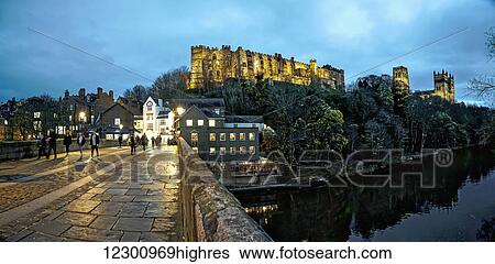 Stock Photograph - Pedestrians walking on a walkway over a river at dusk with Durham Cathedral and Castle on a hilltop; Durham, England. Fotosearch