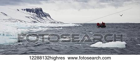 Stock Photograph - People riding in an inflatable boat in the arctic ocean with a view of the snowy cliffs along the coastline; Spitsbergen, Svalbard, Norway. Fotosearch
