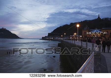 People strolling on the promenade of Passeo de La Concha at dusk, looking towards Mount Urgull with the bay of La Concha to the left; San Sebastian, Spain View Large Photo Image Stock Photo - People strolling on the promenade of Passeo de La Concha at dusk, looking towards Mount Urgull with the bay of La Concha to the left; San Sebastian, Spain. Fotosearch