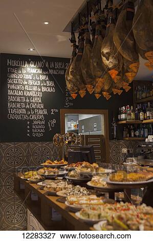 Stock Photo - Pinxos on a bar counter, hams hanging from the ceiling and a menu written on a blackboard in a bar called Portaletas in the old town; San Sebastian, Spain. Fotosearch