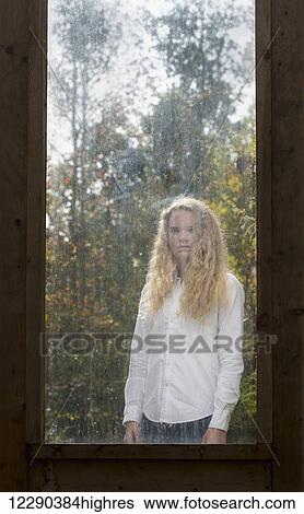 Stock Photograph - Portrait of a caucasian teenage girl with long blond curly hair standing on the other side of a dirty window framed by old wood; Apex, North Carolina, United States of America. Fotosearch