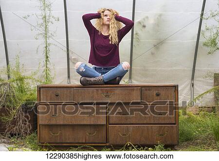 Portrait of a caucasian teenage girl with long blond, curly hair sitting cross-legged on a weathered dresser inside a greenhouse; Apex, North Carolina, United States of America View Large Photo Image Stock Photograph - Portrait of a caucasian teenage girl with long blond, curly hair sitting cross-legged on a weathered dresser inside a greenhouse; Apex, North Carolina, United States of America. Fotosearch