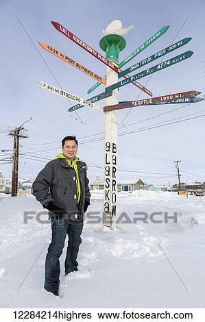 Stock Photograph - Portrait of a man standing in a snowbank beneath the Barrow Milepost, Barrow, North Slope, Arctic Alaska, USA, Winter. Fotosearch