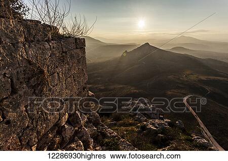 Stock Photograph - Ruins of a stone wall with a sunburst and mountains; Corinth, Greece. Fotosearch