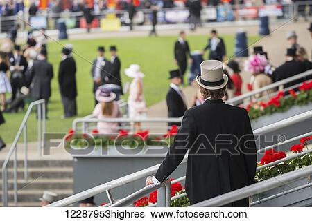 Stock Photograph - Seen from behind in the foreground, a man in black tails and a grey top hat is standing on the steps, looking over at the people in tails and summer dresses on one of the lawns in the Royal Enclosure at Royal Ascot; Ascot, England. Fotosearch