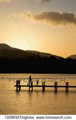 Silhouette of a parent and child walking on a dock on a tranquil lake at sunset; South Lakeland District, England View Large Photo Image Stock Photograph - Silhouette of a parent and child walking on a dock on a tranquil lake at sunset; South Lakeland District, England. Fotosearch