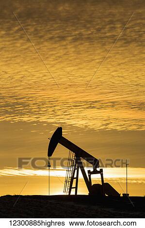 Stock Photograph - Silhouette of a pump jack at sunrise with the sun glowing on the horizon and colourful clouds in the sky; Alberta, Canada. Fotosearch