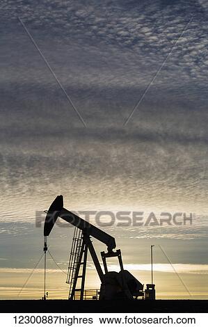 Stock Photograph - Silhouette of a pump jack at sunrise with the sun glowing on the horizon and colourful clouds in the sky; Alberta, Canada. Fotosearch
