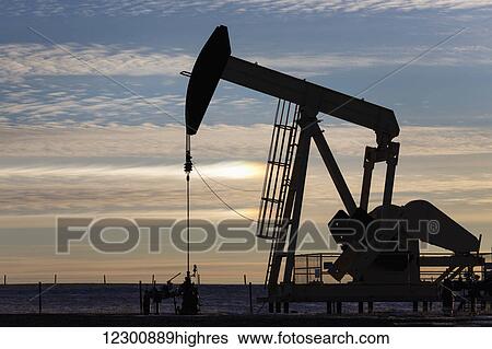 Stock Photograph - Silhouette of a pump jack at sunrise with the sun glowing on the horizon and colourful clouds in the sky; Alberta, Canada. Fotosearch