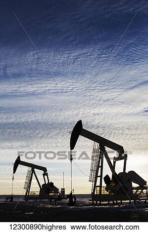 Stock Photograph - Silhouette of a pump jack at sunrise with the sun glowing on the horizon and colourful clouds in the sky; Alberta, Canada. Fotosearch