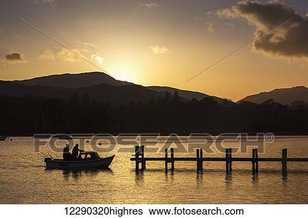 Stock Photograph - Silhouette of fishermen in a boat arriving at a dock on a lake at sunset; Keswick, Cumbria, England. Fotosearch