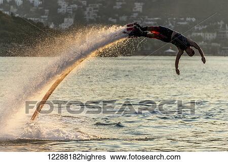 Stock Photograph - Silhouette of Flyboarder diving in perfect high backlit arc in the late afternoon sunshine with a village in the background; Torba, Mugla Province, Turkey. Fotosearch