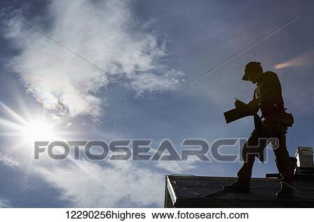 Stock Photograph - Silhouette of male roofer on top of roof holding new shingles with clouds sun burst and blue sky; Calgary, Alberta, Canada. Fotosearch