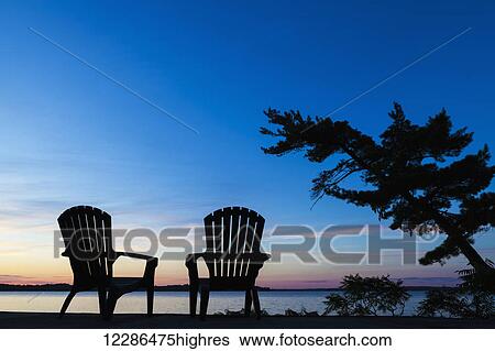 Stock Photograph - Silhouette of Muskoka chairs and Balsam Lake at sunrise; Ontario, Canada. Fotosearch