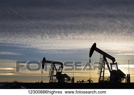 Stock Photograph - Silhouette of two pump jacks at sunrise with the sun glowing on the horizon and colourful clouds in the sky; Alberta, Canada. Fotosearch