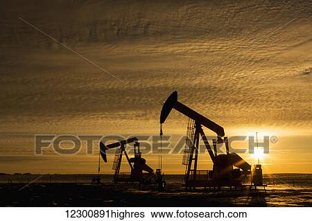 Stock Photograph - Silhouette of two pump jacks at sunrise with sun burst and colourful clouds in the sky; Alberta, Canada. Fotosearch