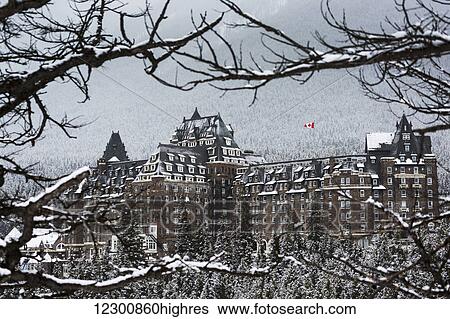 Stock Photograph - Snow covered Banff Springs Hotel framed by tree limbs with snow covered trees on mountain hillside in the background; Banff, Alberta, Canada. Fotosearch