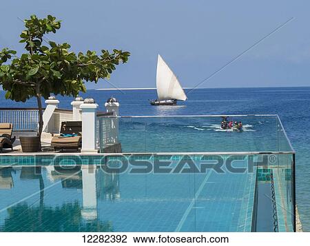 Stock Image - Swimming pool of a resort at the edge of the ocean with a dhow in the water; Zanzibar, Tanzania. Fotosearch