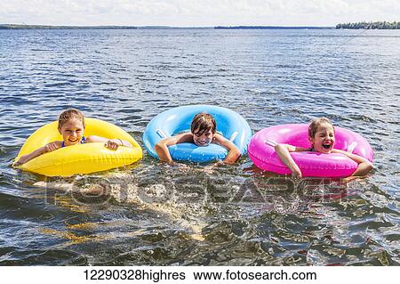 Stock Photograph - Three kids playing in Balsam Lake wearing colorful inflatable rings; Ontario, Canada. Fotosearch
