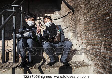 Stock Photograph - Two Asian tourists with masks sitting at the Colosseum; Rome, Italy. Fotosearch