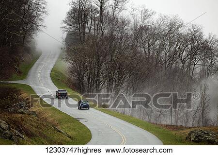 Stock Photograph - Two cars traveling on a rainy, winding Blue Ridge Parkway with a fog and mist hiding the road in front of them in early spring with no leaves on the trees; North Carolina, United States of America. Fotosearch
